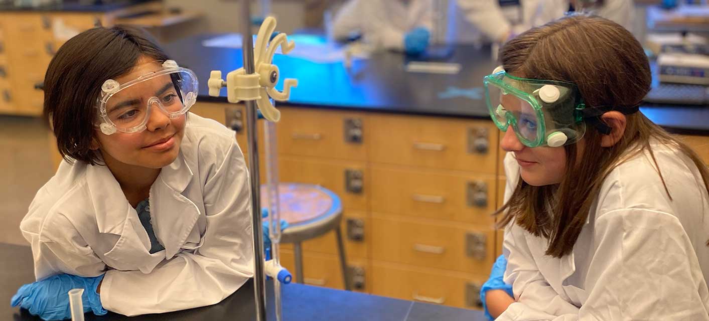 Two girls wearing a lab coat, gloves, and goggles